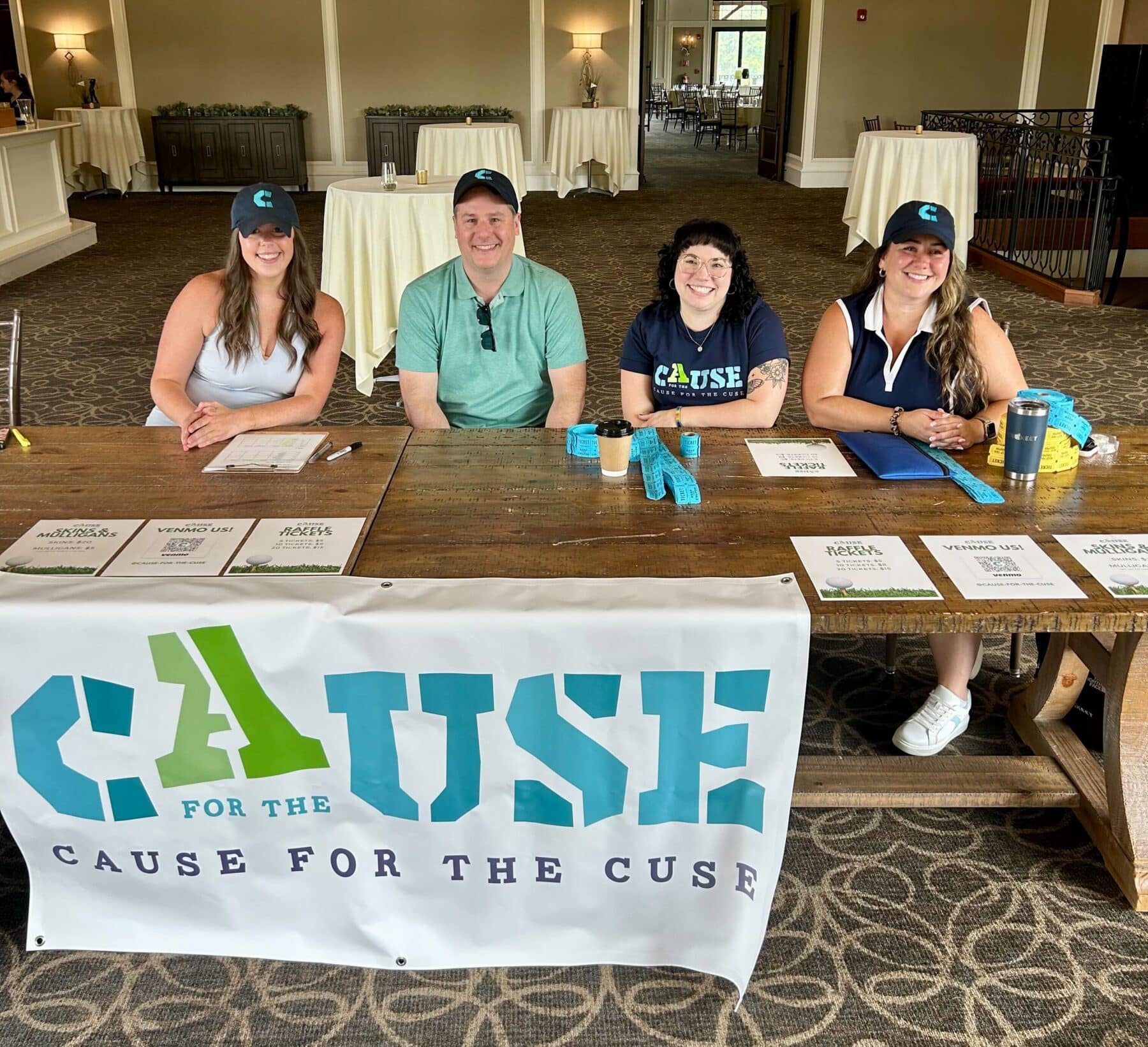 Four Cause For The Cuse volunteers sit at the tournament sign up table, smiling.